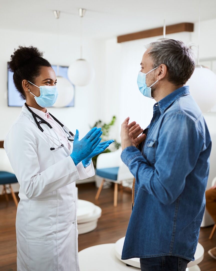 Happy African American doctor communicating with her patient in waiting room while wearing face masks due to coronavirus pandemic.